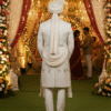 A man in a cream-colored sherwani with intricate embroidery, a matching pagri (turban), and a pearl necklace stands in front of a wedding arch decorated with flowers.