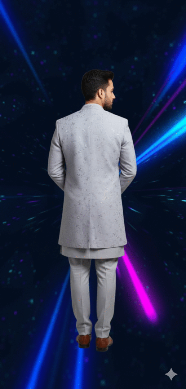 A man wearing a grey embroidered indo-western sherwani set, with a long jacket over a kurta and pants, standing against a dark background with blue and pink light streaks.