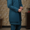 A man wearing a teal blue Indo-Western suit with a long jacket and matching pants, standing indoors.
