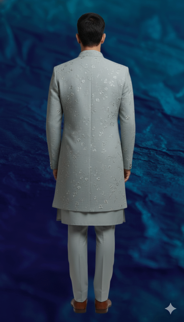 A man stands against a textured blue background, wearing a dove grey Indo-Western sherwani with intricate embroidery, paired with a matching kurta and trousers.