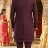 A man wearing a wine-colored embroidered Indo-Western sherwani set with a long jacket, standing in a traditional Indian architectural setting.