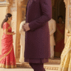 A man wearing a wine-colored embroidered Indo-Western sherwani set with a long jacket, standing in a traditional Indian architectural setting.