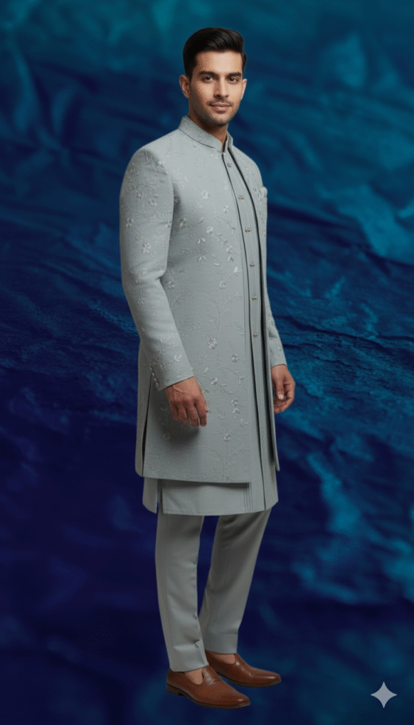 A man stands against a textured blue background, wearing a dove grey Indo-Western sherwani with intricate embroidery, paired with a matching kurta and trousers.