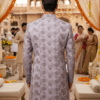 A man wearing a lavender floral embroidered sherwani with a mandarin collar, standing in a traditional wedding setting decorated with marigold garlands.