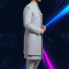 A man wearing a grey embroidered indo-western sherwani set, with a long jacket over a kurta and pants, standing against a dark background with blue and pink light streaks.