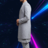 A man wearing a grey embroidered indo-western sherwani set, with a long jacket over a kurta and pants, standing against a dark background with blue and pink light streaks.