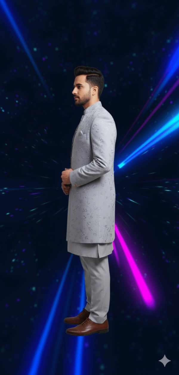 A man wearing a grey embroidered indo-western sherwani set, with a long jacket over a kurta and pants, standing against a dark background with blue and pink light streaks.