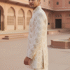 Cream-colored Indo-Western suit for men featuring a long embroidered jacket with golden floral patterns, worn with matching kurta and trousers in a heritage palace setting.