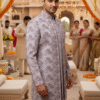 A man wearing a lavender floral embroidered sherwani with a mandarin collar, standing in a traditional wedding setting decorated with marigold garlands.