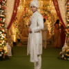 A man in a cream-colored sherwani with intricate embroidery, a matching pagri (turban), and a pearl necklace stands in front of a wedding arch decorated with flowers.