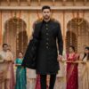 A man wearing a black brocade Indo-Western sherwani with gold buttons, standing confidently in a traditional palace courtyard decorated with marigold garlands and lights.