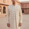 Cream-colored Indo-Western suit for men featuring a long embroidered jacket with golden floral patterns, worn with matching kurta and trousers in a heritage palace setting.