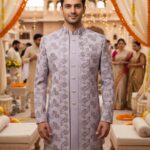 A man wearing a lavender floral embroidered sherwani with a mandarin collar, standing in a traditional wedding setting decorated with marigold garlands.