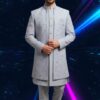 A man wearing a grey embroidered indo-western sherwani set, with a long jacket over a kurta and pants, standing against a dark background with blue and pink light streaks.