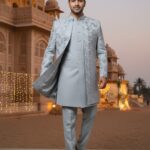 A man wearing a silver Indo-Western sherwani with intricate embroidery stands in front of a palace-like building at dusk.