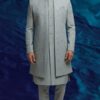 A man stands against a textured blue background, wearing a dove grey Indo-Western sherwani with intricate embroidery, paired with a matching kurta and trousers.