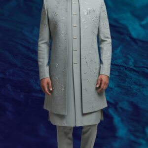 A man stands against a textured blue background, wearing a dove grey Indo-Western sherwani with intricate embroidery, paired with a matching kurta and trousers.