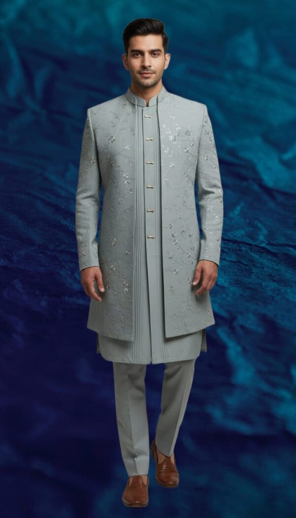 A man stands against a textured blue background, wearing a dove grey Indo-Western sherwani with intricate embroidery, paired with a matching kurta and trousers.