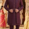 A man wearing a wine-colored embroidered Indo-Western sherwani set with a long jacket, standing in a traditional Indian architectural setting.