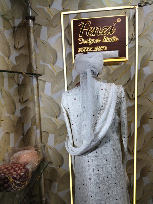 Elegant white sherwani with intricate embroidery, pearl mala, and turban displayed at a designer studio.