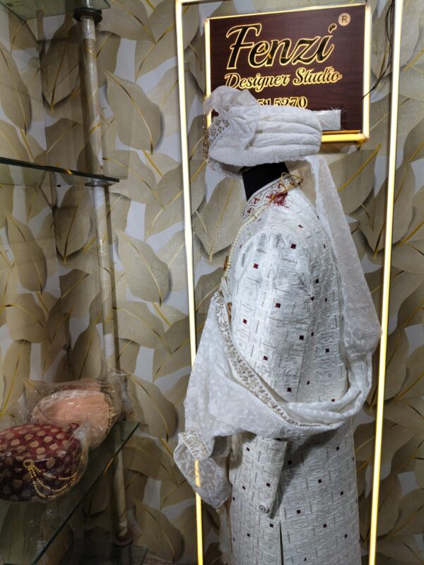 Elegant white sherwani with intricate embroidery, pearl mala, and turban displayed at a designer studio.