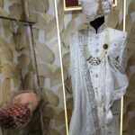 Elegant white sherwani with intricate embroidery, pearl mala, and turban displayed at a designer studio.