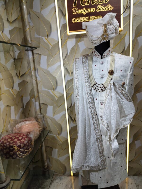 Elegant white sherwani with intricate embroidery, pearl mala, and turban displayed at a designer studio.